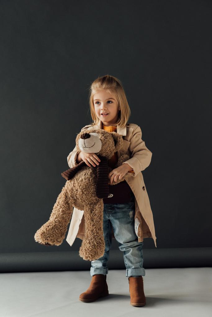 smiling and cute child in trench coat and jeans holding teddy bear on black background  - Photo, Image