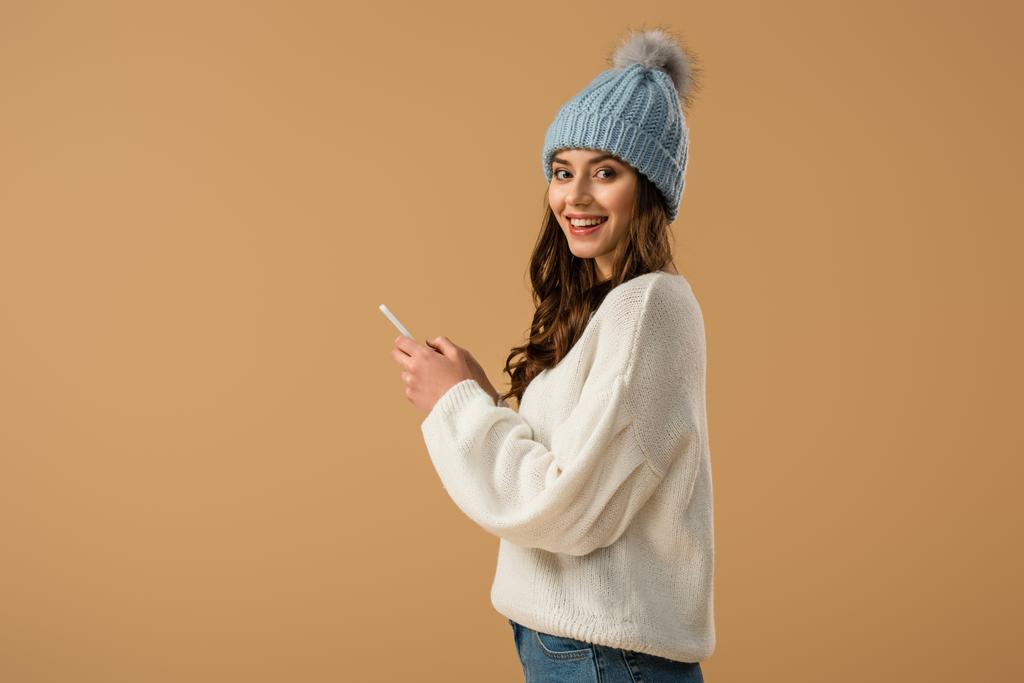 Excited curly woman in knitted hat holding smartphone isolated on beige - Photo, Image