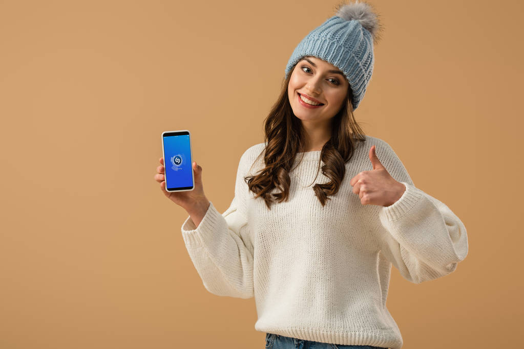 Chica morena feliz sosteniendo teléfono inteligente con aplicación shazam en la pantalla y mostrando el pulgar hacia arriba aislado en beige
 - Foto, imagen