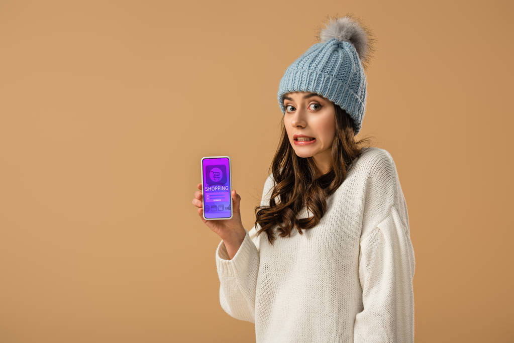 Chica morena confusa en sombrero sosteniendo smartphone con aplicación de compras en pantalla aislada en beige
 - Foto, Imagen