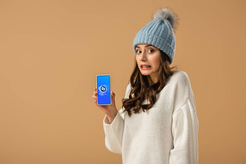 Mujer joven confundida sosteniendo teléfono inteligente con aplicación shazam en la pantalla aislada en beige
 - Foto, imagen