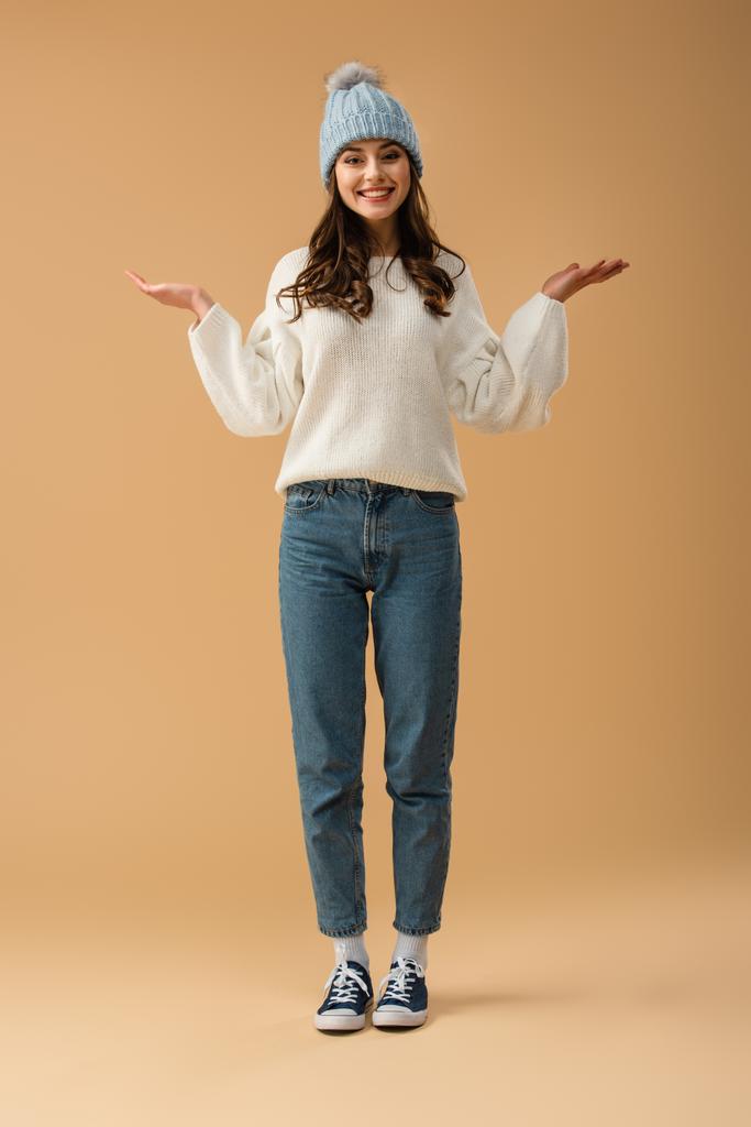 Ganzkörperansicht des lachenden brünetten Mädchens in Jeans und Pullover auf beigem Hintergrund - Foto, Bild