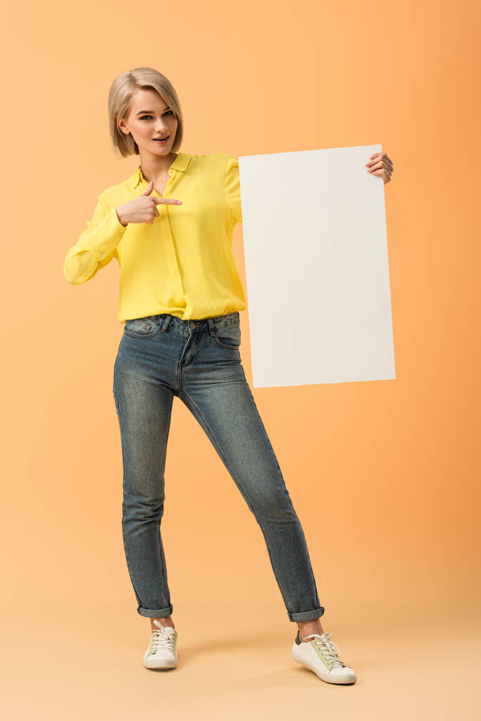 selbstbewusstes blondes Mädchen in Jeans zeigt mit dem Finger auf leeres Plakat auf orangefarbenem Hintergrund - Foto, Bild