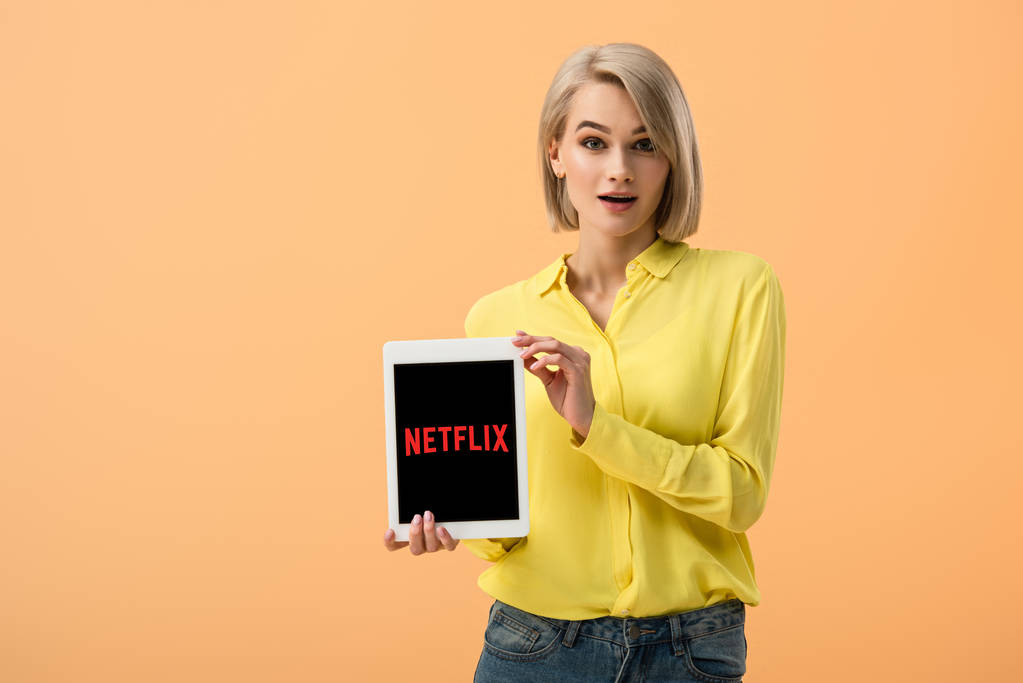 Удивлена стильная женщина в желтой рубашке с цифровым планшетом с netflix приложение на экране изолированы на оранжевый
 - Фото, изображение
