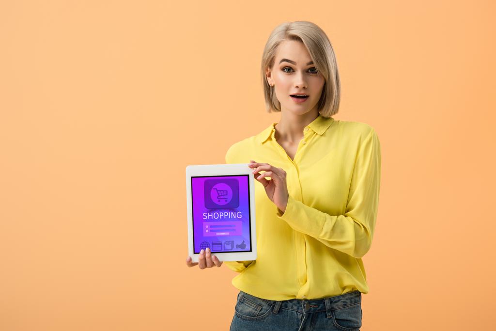 Έκπληκτος ξανθιά γυναίκα κρατώντας ψηφιακή δισκίο με online αγορές app στην οθόνη που απομονώνονται σε πορτοκαλί - Φωτογραφία, εικόνα
