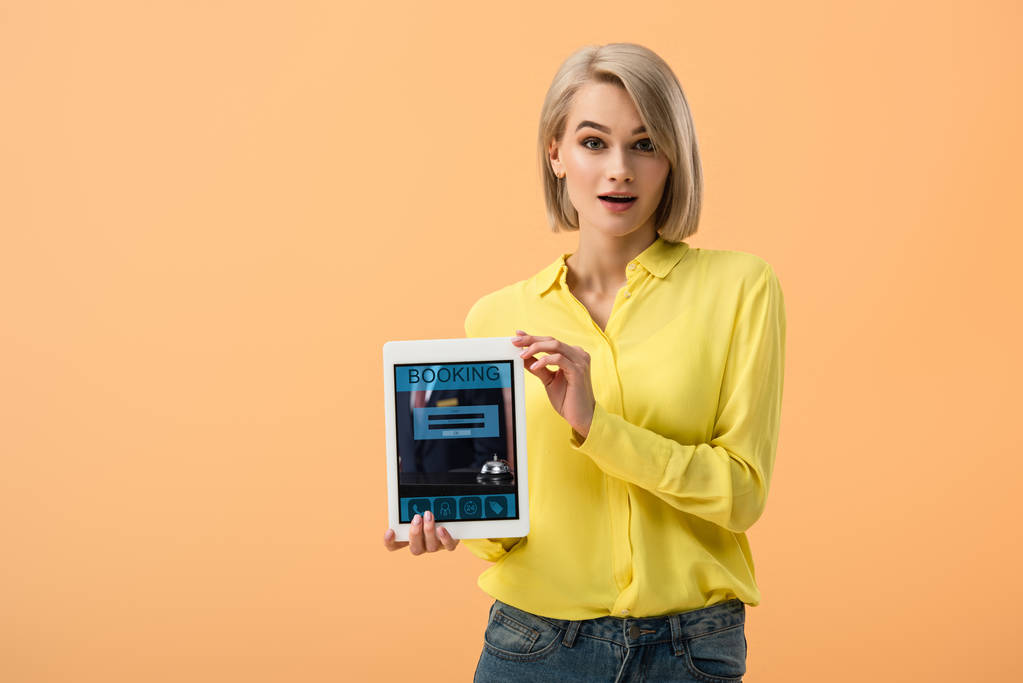 Überraschtes Mädchen im gelben Hemd hält digitales Tablet mit Buchungs-App auf dem Bildschirm isoliert auf orange - Foto, Bild