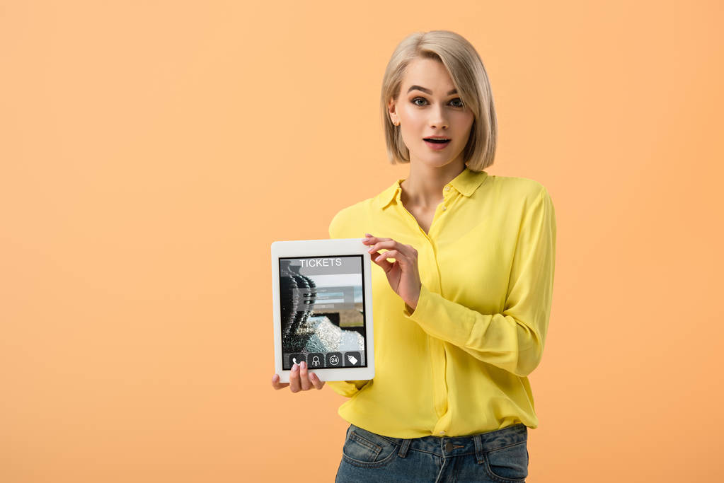 Mujer rubia sorprendida sosteniendo tableta digital con tickets app en pantalla aislada en naranja
 - Foto, Imagen