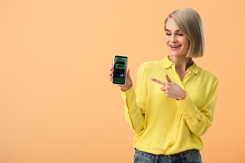 Smiling woman in yellow shirt pointing with finger at smartphone with booking app on screen isolated on orange - Photo, Image