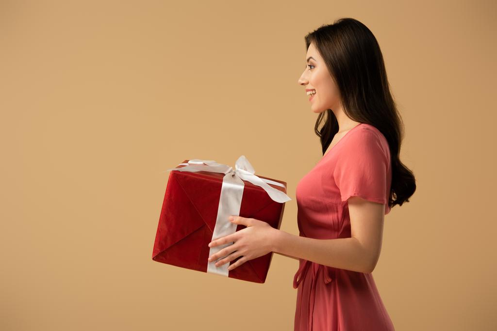 Вид сбоку счастливой брюнетки в платье с подарочной коробкой, изолированной на бежевом
 - Фото, изображение