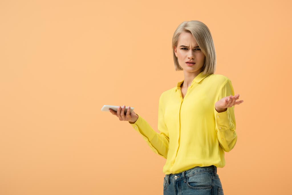 Недовольная блондинка в жёлтой рубашке держит смартфон изолированным на оранжевом
 - Фото, изображение