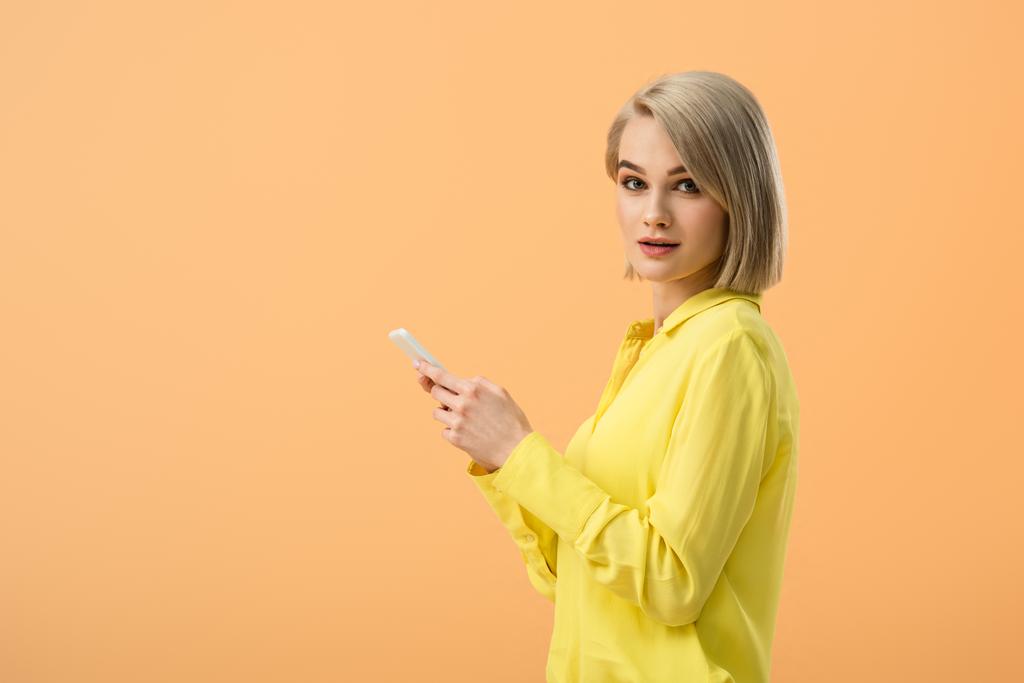 Mujer rubia bonita en camisa amarilla sosteniendo teléfono inteligente y mirando a la cámara aislada en naranja
 - Foto, imagen