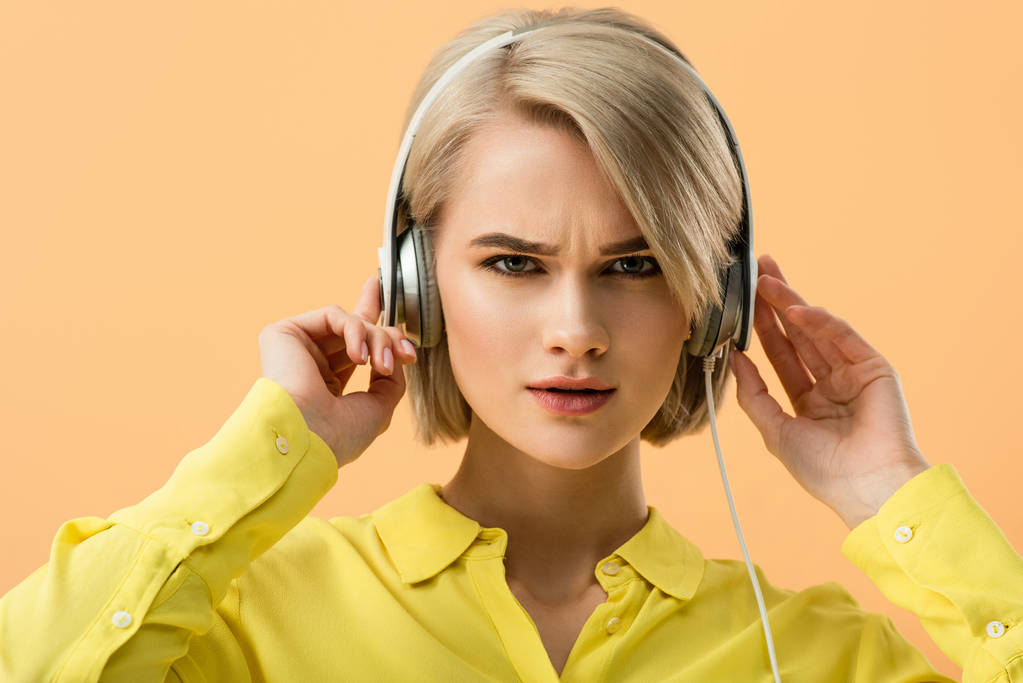 Δυσαρεστημένοι ξανθιά νεαρή γυναίκα αγγίζοντας ακουστικά και βλέπουν τα φωτογραφικών μηχανών που απομονώνονται σε πορτοκαλί - Φωτογραφία, εικόνα