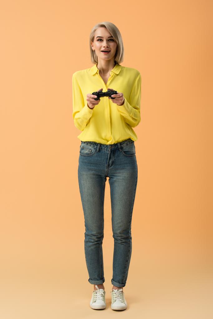 Visão de comprimento total da menina loira feliz em jeans segurando joystick no fundo laranja
 - Foto, Imagem