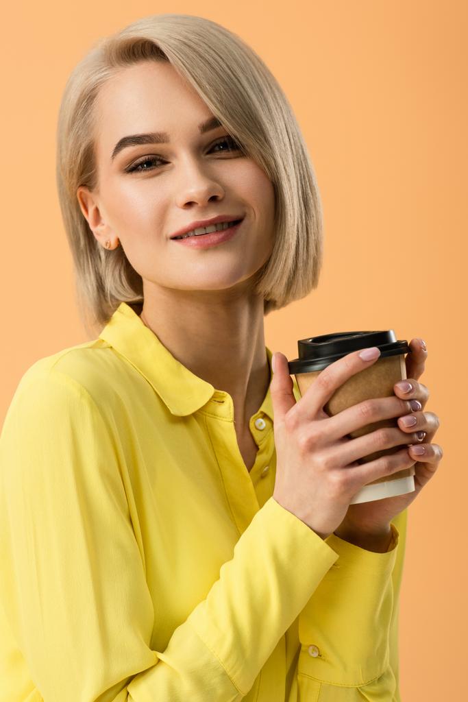 Χαμογελώντας όμορφη ξανθιά κοπέλα κρατώντας χαρτί Κύπελλο του καφέ που απομονώνονται σε πορτοκαλί - Φωτογραφία, εικόνα