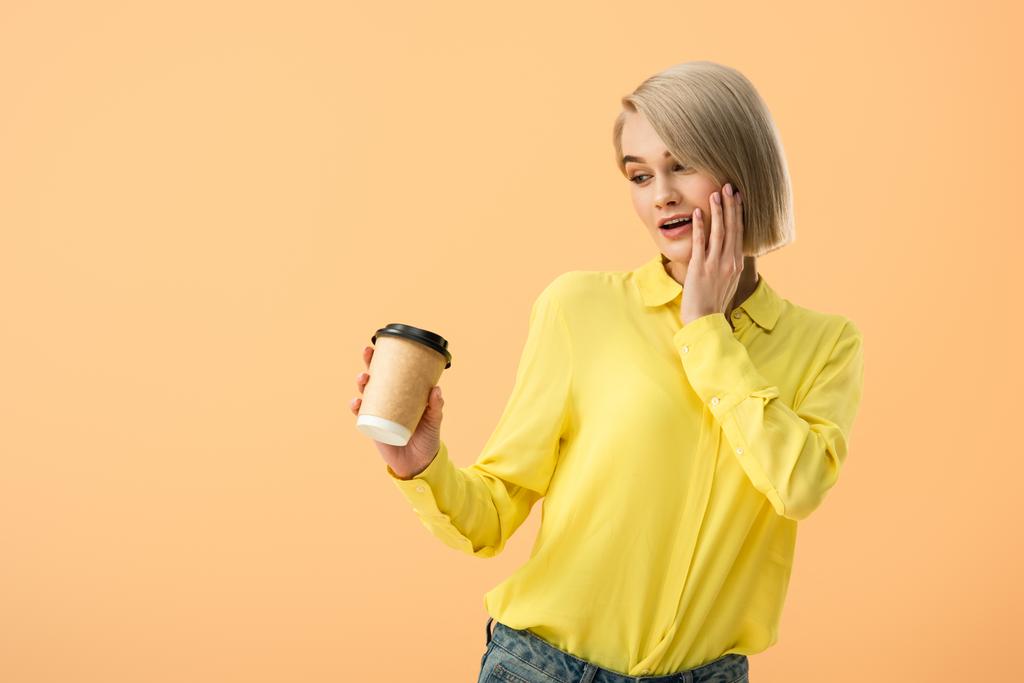 Заинтересованная молодая женщина в желтой рубашке, глядя на бумажную чашку с кофе изолированы на оранжевый
 - Фото, изображение