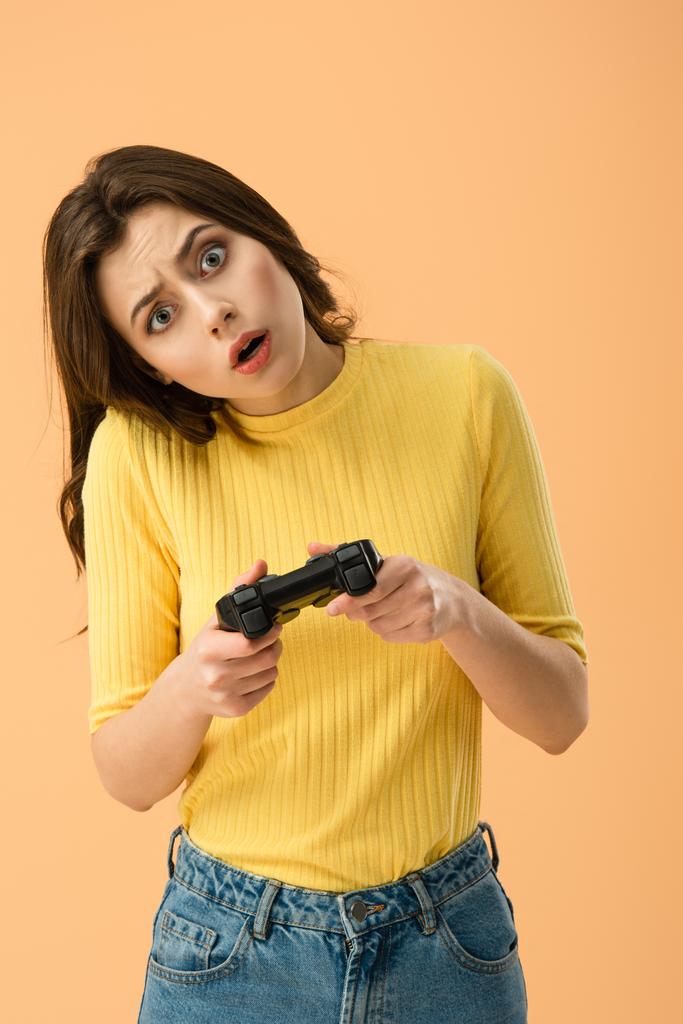 Zszokowany brunetka, młoda kobieta trzyma joystick na pomarańczowym tle - Zdjęcie, obraz