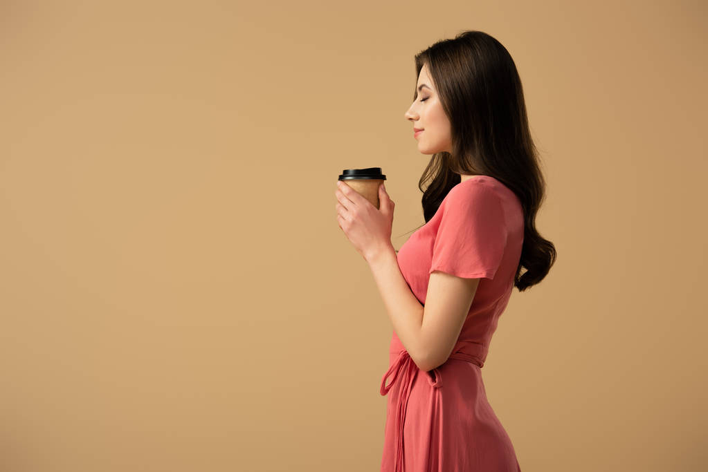 ベージュに分離されたコーヒーのカップを保持しているエレガントなブルネットの女性の側面図 - 写真・画像