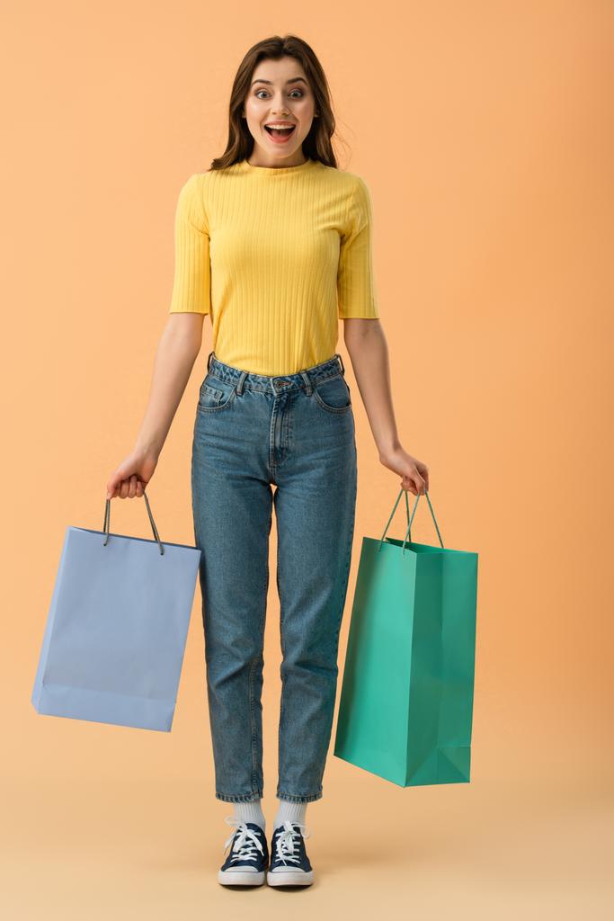 Volle lengte weergave van verrast lachende brunette meisje oranje achtergrond boodschappentassen vasthouden - Foto, afbeelding
