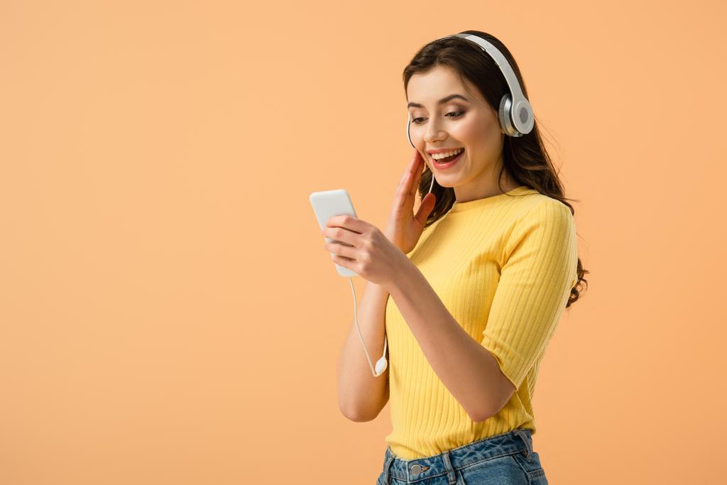 Chica morena alegre en auriculares con teléfono inteligente con sonrisa aislada en naranja
 - Foto, imagen