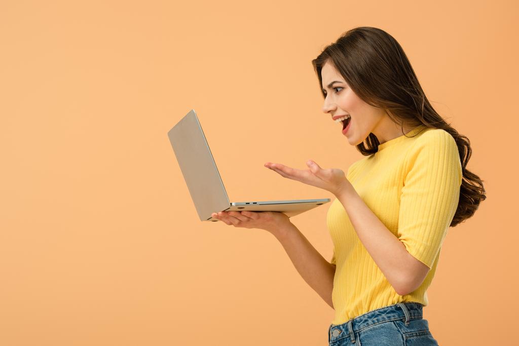 Συναισθηματική μελαχρινή νεαρή γυναίκα κρατώντας το laptop που απομονώνονται σε πορτοκαλί - Φωτογραφία, εικόνα
