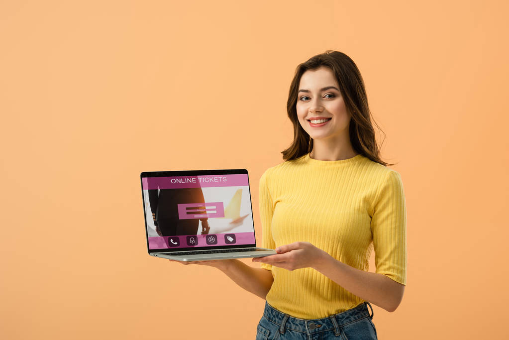 Mosolygós barna lány gazdaság a Jegymester honlapján laptop képernyőn elszigetelt narancs - Fotó, kép