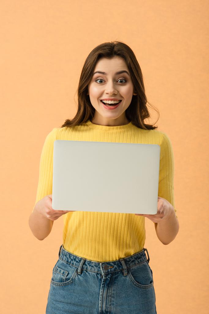 Здивована брюнетка дівчина тримає ноутбук і посміхається ізольовано на помаранчевому
 - Фото, зображення