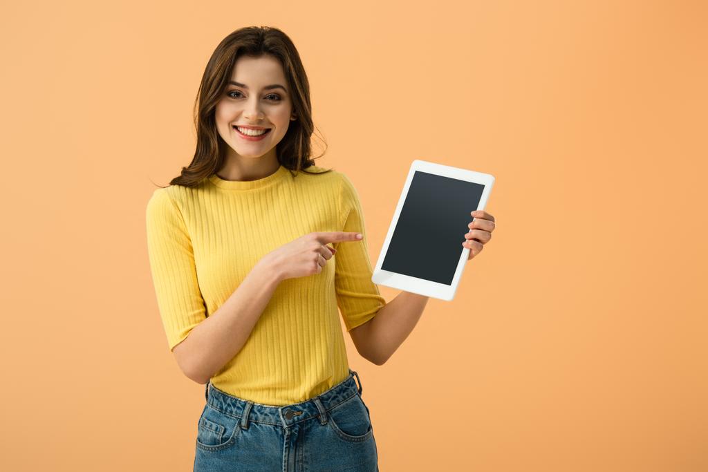 Hymyilevä brunette tyttö osoittaa sormella digitaalinen tabletti tyhjä näyttö eristetty oranssi
 - Valokuva, kuva