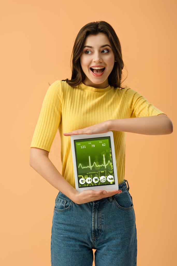 Радостная брюнетка женщина держит цифровой планшет с приложением здоровья на экране изолированы на оранжевый
 - Фото, изображение
