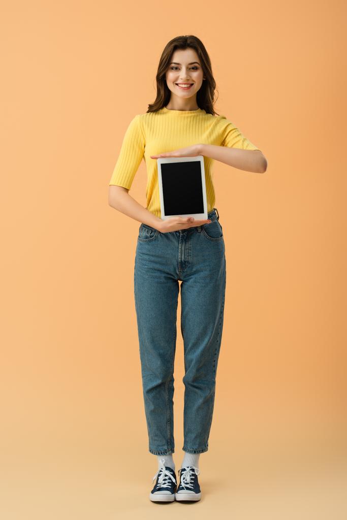 オレンジ色の背景の空白の画面とデジタル タブレットを保持しているジーンズで微笑んでいる女の子の完全な長さのビュー - 写真・画像
