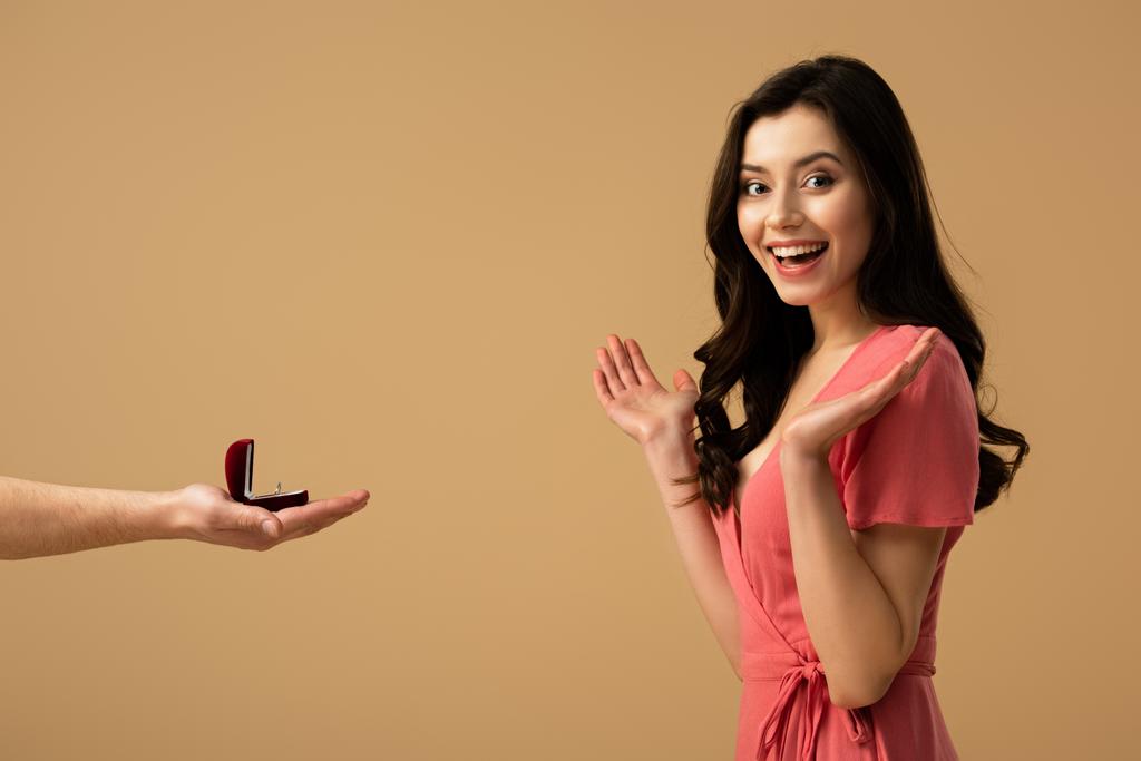 Περικοπεί θεώρηση του ανθρώπου που κρατώντας δαχτυλίδι και να κάνει πρόταση γάμου ενθουσιασμένος μελαχρινή κοπέλα που απομονώνονται σε μπεζ - Φωτογραφία, εικόνα