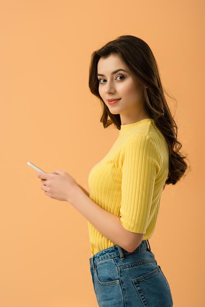 Krásná bruneta dívka drží smartphone a při pohledu na fotoaparát izolované na oranžové - Fotografie, Obrázek