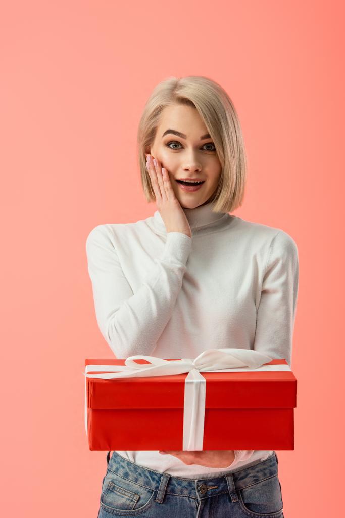 schockierte blonde Frau hält rote Geschenkschachtel isoliert auf rosa  - Foto, Bild