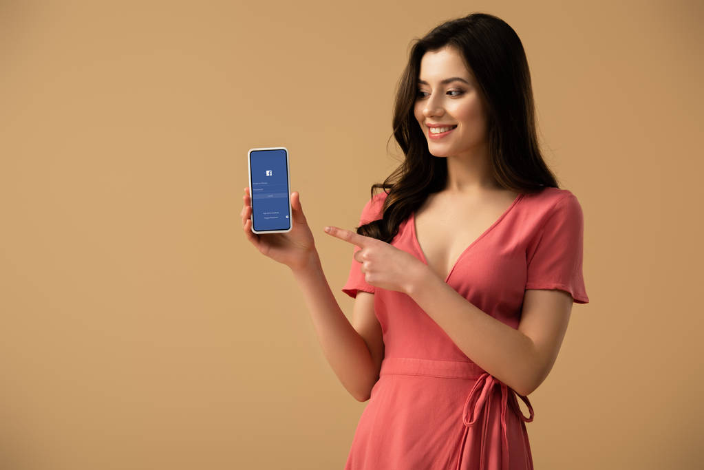 joyeuse fille brune pointant du doigt le smartphone avec application facebook à l'écran isolé sur brun
  - Photo, image