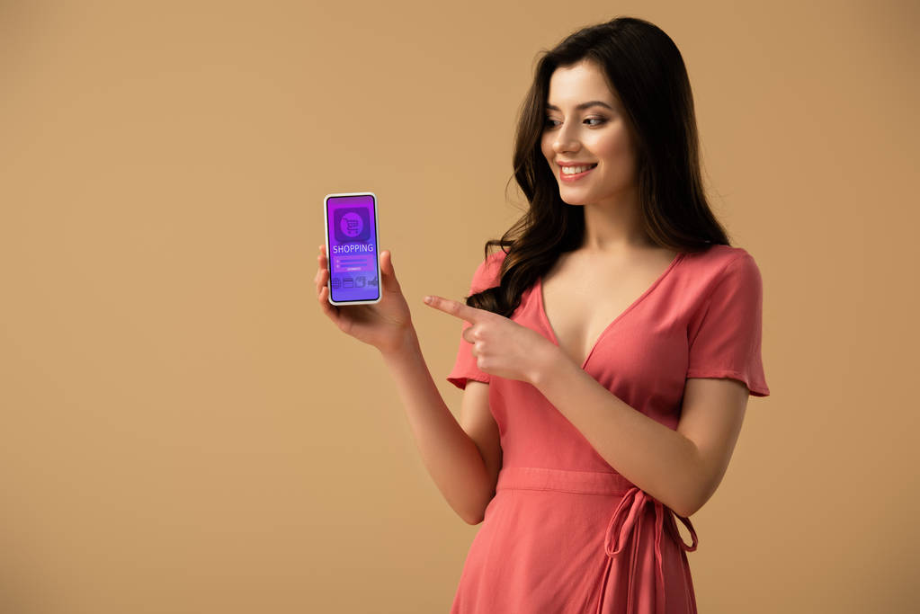 alegre morena chica apuntando con el dedo en el teléfono inteligente con la aplicación de compras en la pantalla aislado en marrón
  - Foto, Imagen