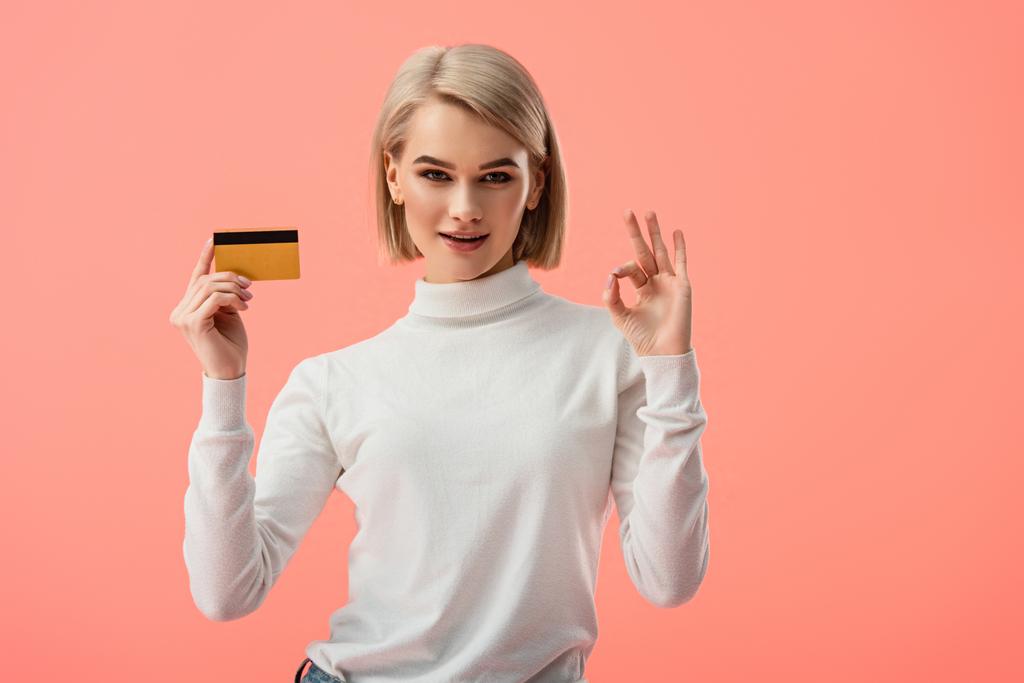 щаслива блондинка молода жінка тримає кредитну картку і показує знак ОК ізольовано на рожевому
  - Фото, зображення