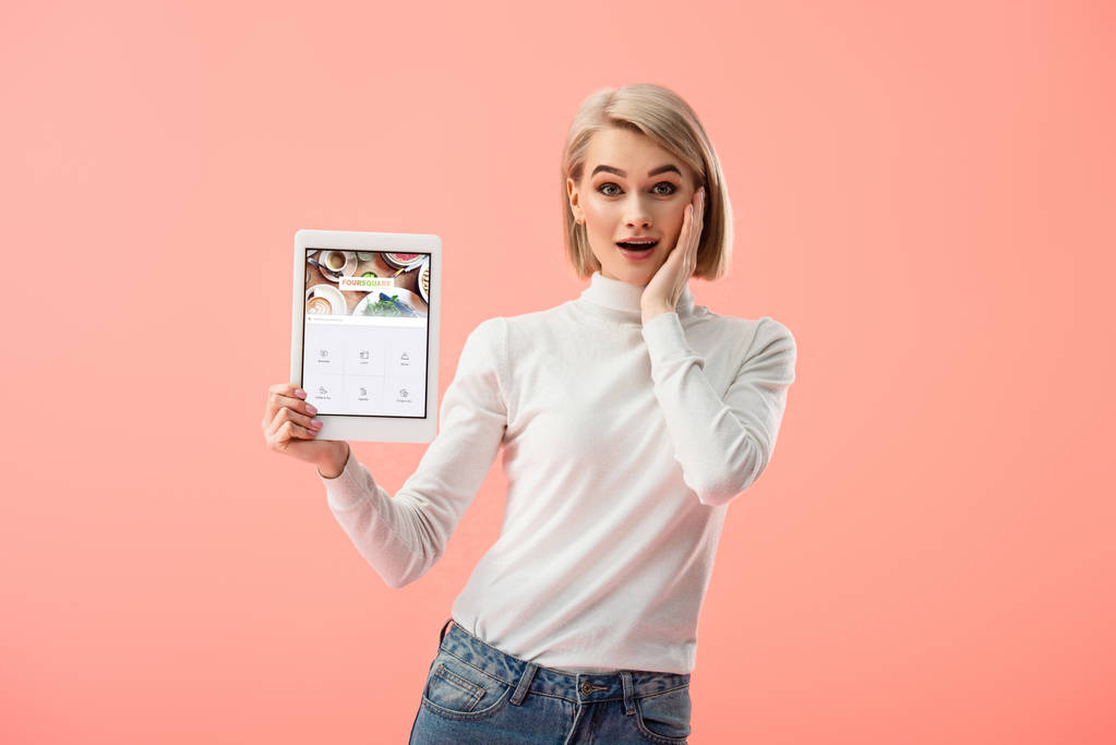 zaskoczony Kobieta blonde holding cyfrowego tabletu z foursquare aplikacji na ekranie na różowym tle  - Zdjęcie, obraz