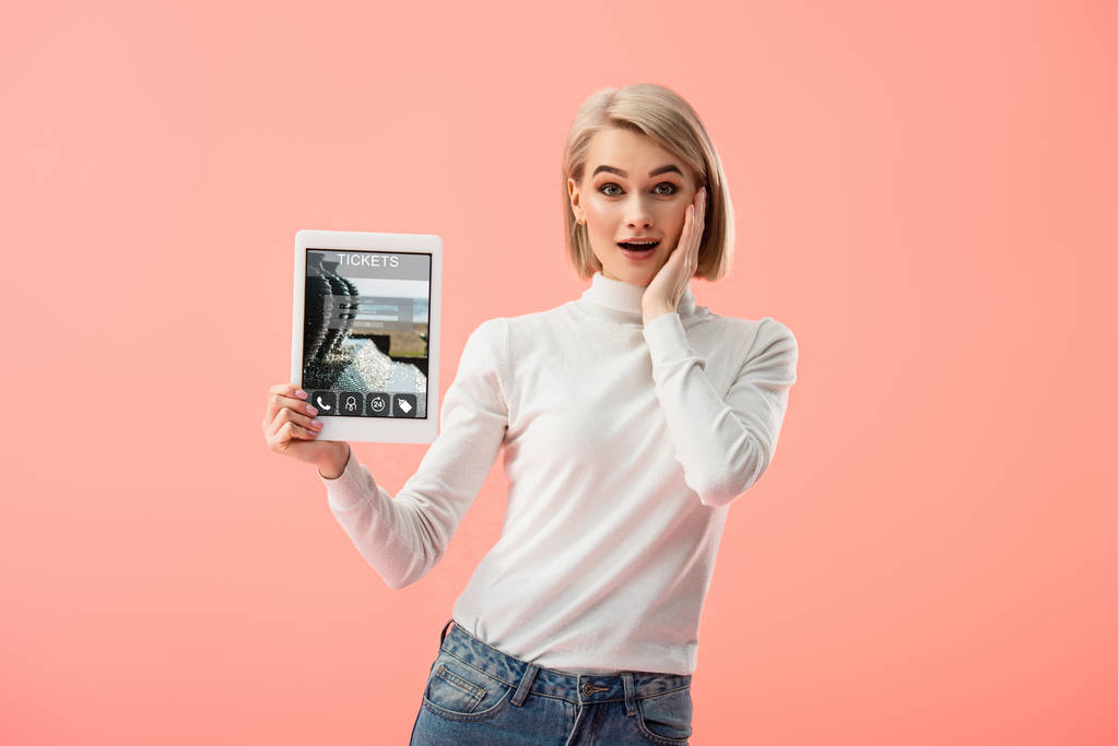 удивленный блондинка держит цифровой планшет с билетами приложение на экране изолированы на розовый
  - Фото, изображение