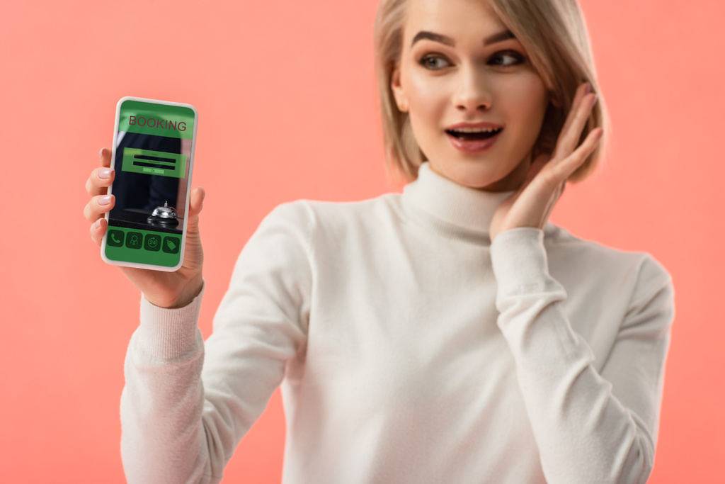 selective focus of surprised blonde woman holding smartphone with booking app on screen isolated on pink  - Photo, Image
