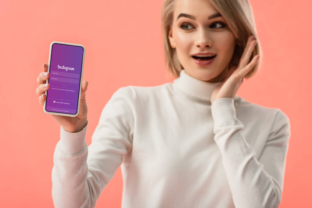 Selektiver Fokus einer überraschten blonden Frau, die ihr Smartphone mit Instagram-App auf dem Bildschirm isoliert auf rosa hält  - Foto, Bild