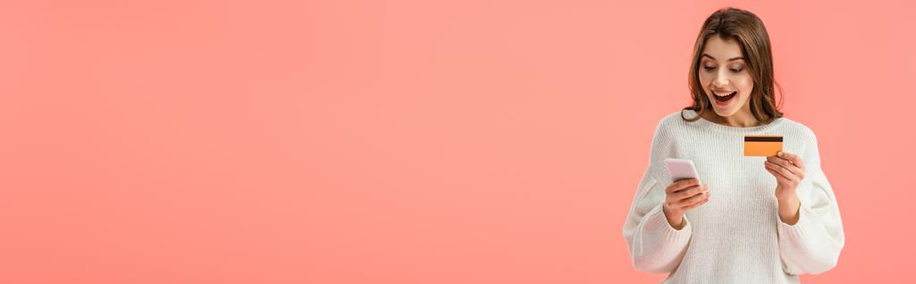 Panoramaaufnahme eines überraschten brünetten Mädchens mit Kreditkarte und Smartphone isoliert auf rosa - Foto, Bild