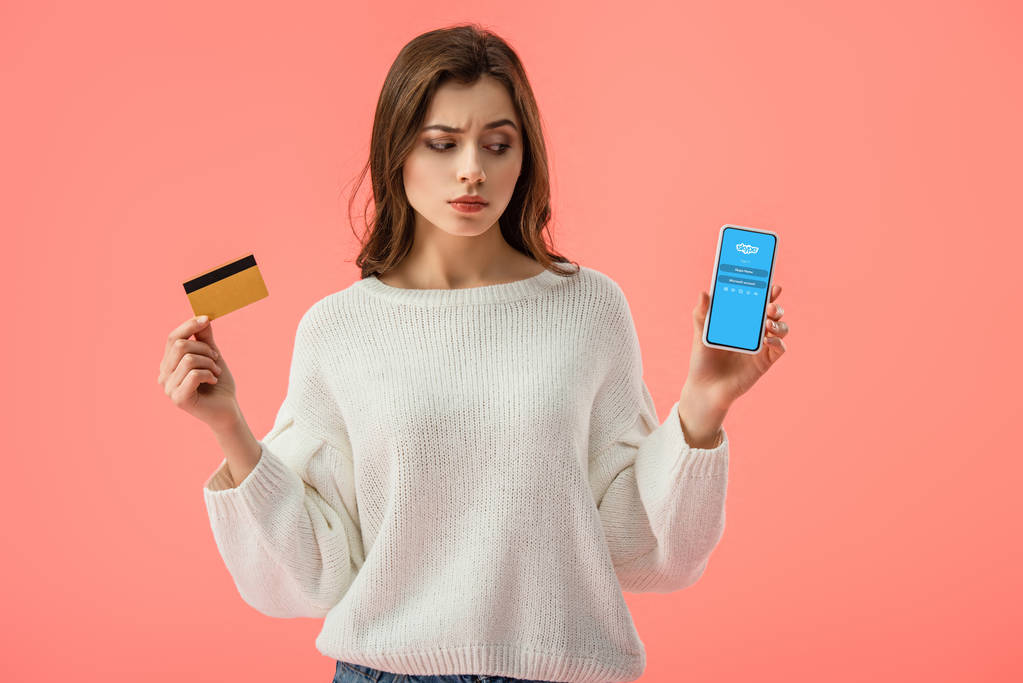 atractiva chica morena sosteniendo tarjeta de crédito mientras mira el teléfono inteligente con aplicación skype en la pantalla aislada en rosa
  - Foto, imagen