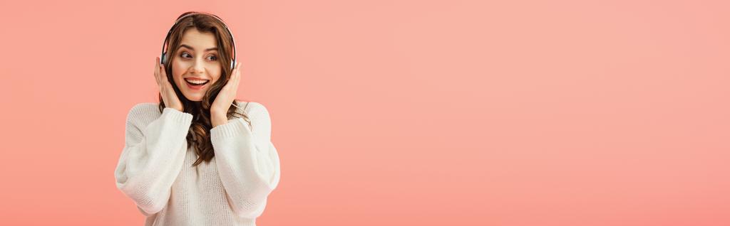πανοραμικό στιγμιότυπο της χαμογελώντας και όμορφη γυναίκα στο λευκό πουλόβερ ακούτε μουσική με ακουστικά που απομονώνονται σε ροζ - Φωτογραφία, εικόνα