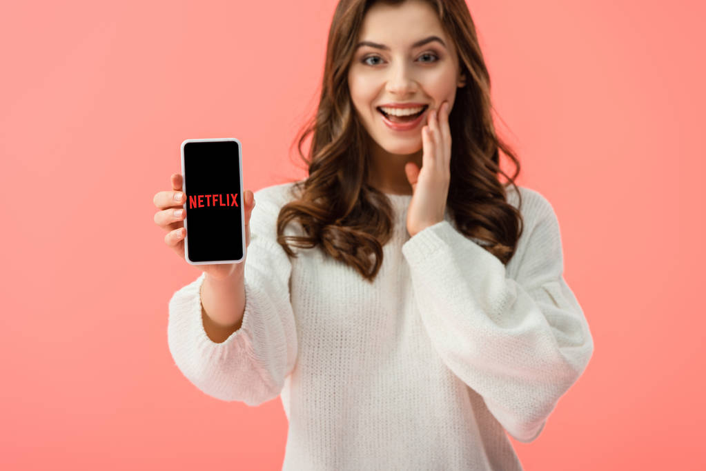 επιλεκτική εστίαση γυναίκα στο λευκό πουλόβερ κρατώντας smartphone με εφαρμογή netflix στην οθόνη που απομονώνονται σε ροζ  - Φωτογραφία, εικόνα