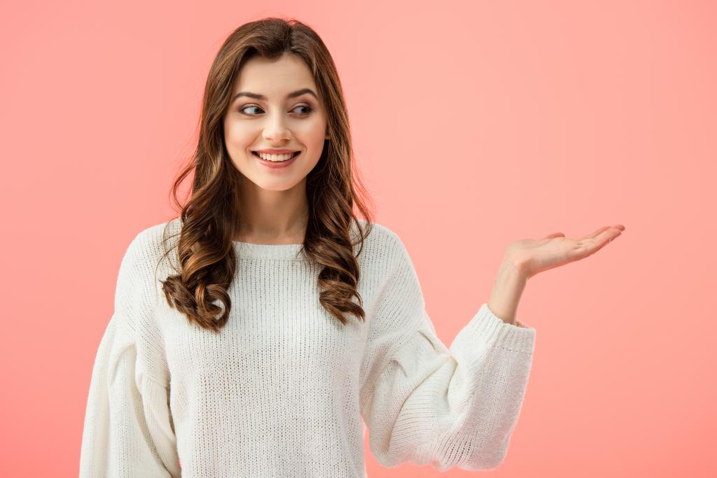 smiling and beautiful woman in white sweater pointing with hand isolated on pink - Photo, Image