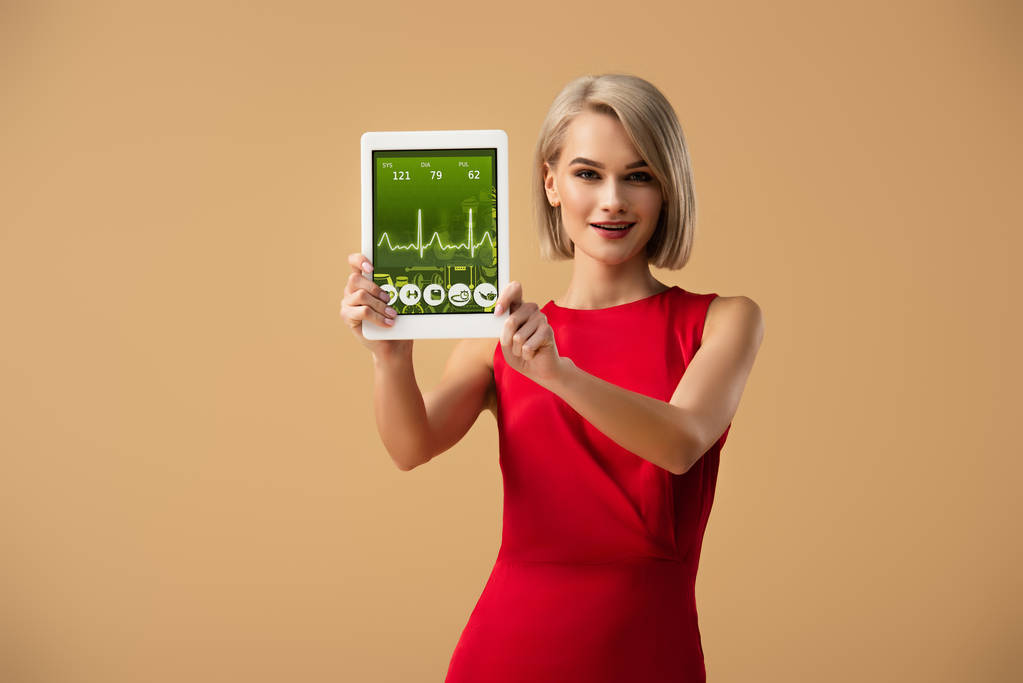 piękna kobieta w czerwonej sukience gospodarstwa cyfrowego tabletu z aplikacji Zdrowie na ekranie na beżowym tle - Zdjęcie, obraz