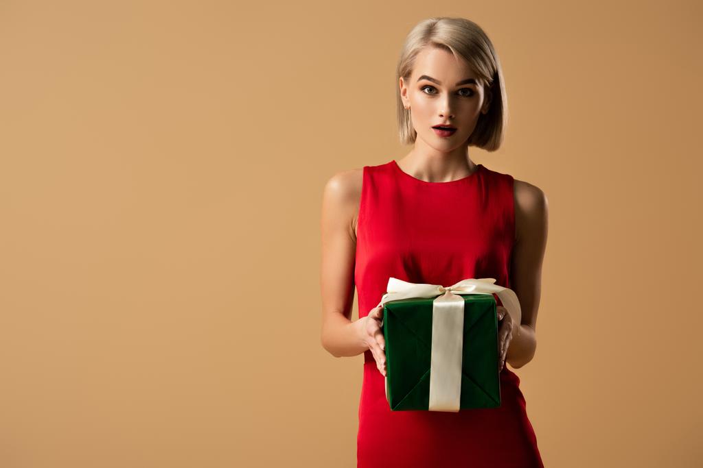 surprised and beautiful young woman in red dress holding gift box isolated on beige - Photo, Image
