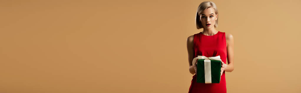 панорамний знімок здивованої і красивої молодої жінки в червоній сукні, що тримає подарункову коробку ізольовано на бежевому
 - Фото, зображення