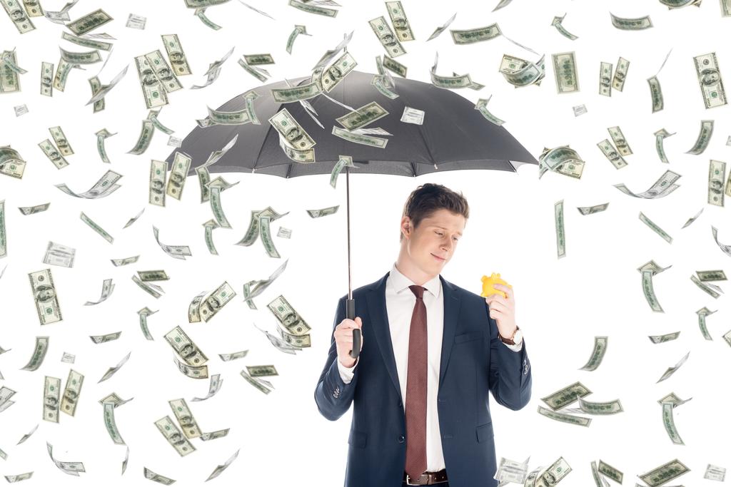 επιτυχημένος επιχειρηματίας με κοστούμι κρατώντας ομπρέλα και κίτρινο κουμπαράς υπό βροχή χρήματα σε λευκό φόντο - Φωτογραφία, εικόνα