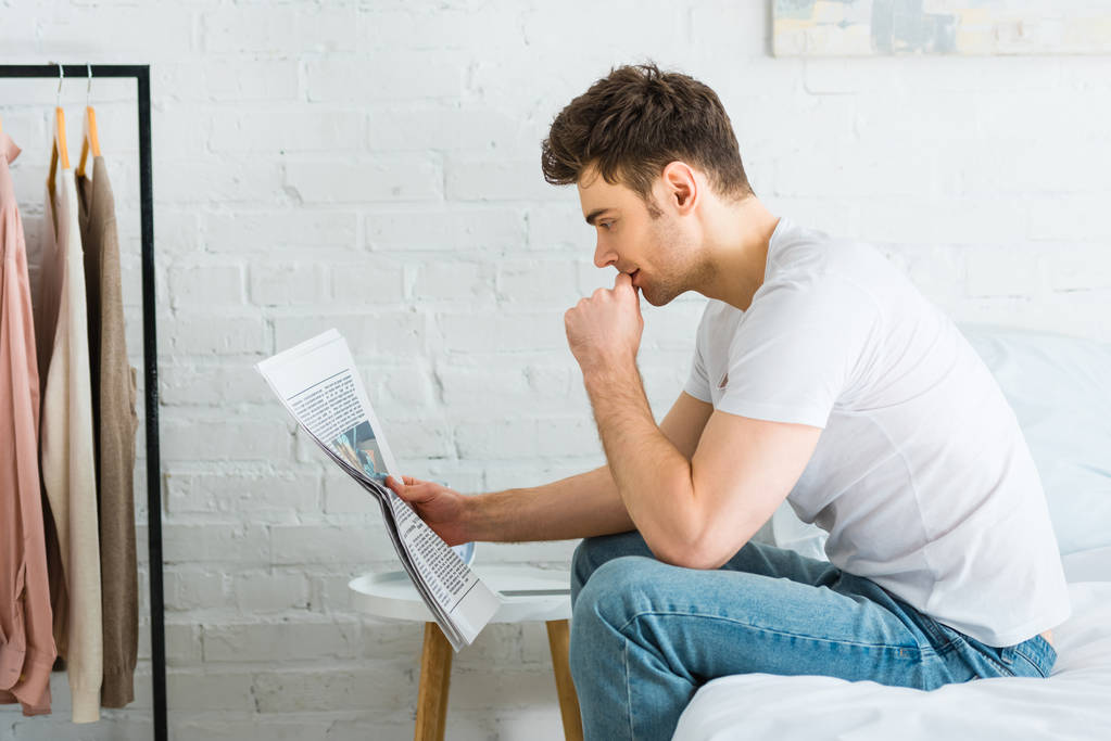 Beyaz t-shirt ve jeans yatağın üstüne oturan ve evde gazete okuyan adam - Fotoğraf, Görsel