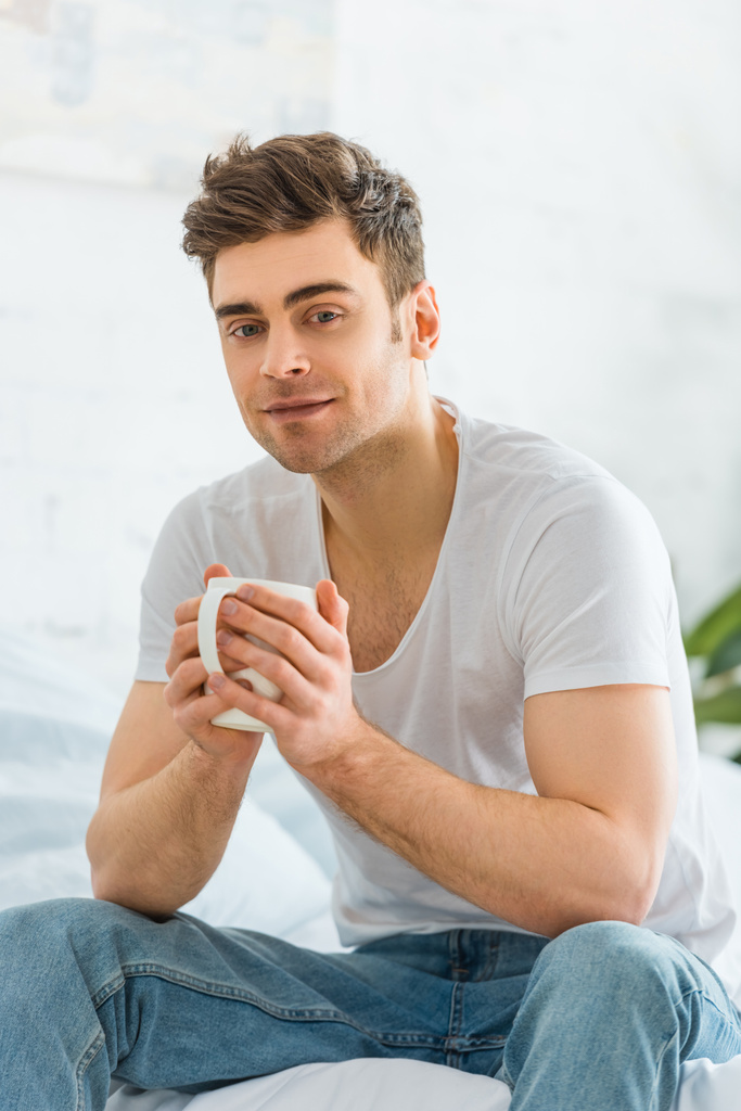 όμορφος άνδρας στο λευκό t-shirt και τζιν που κάθεται στο κρεβάτι με το κύπελλο στο υπνοδωμάτιο - Φωτογραφία, εικόνα